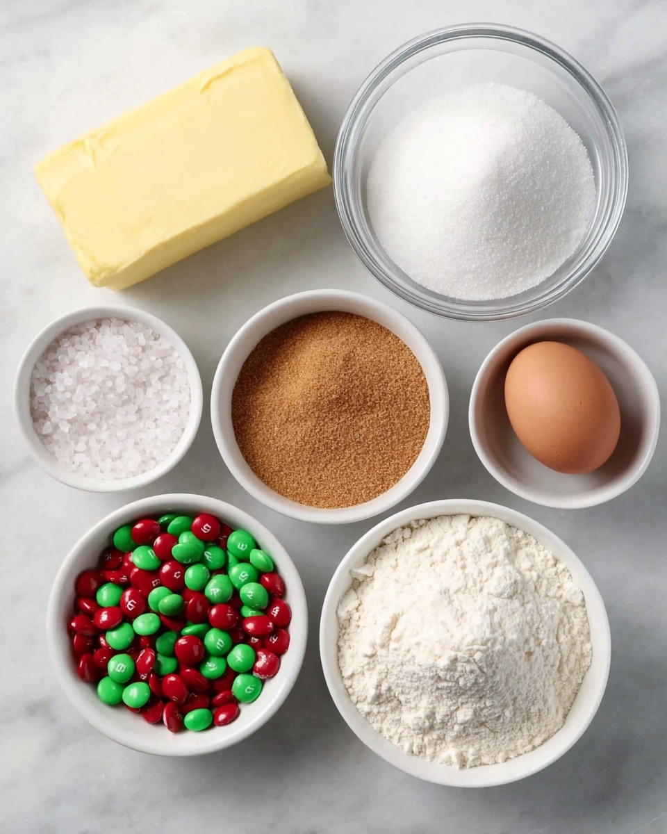 Christmas M&M Cookies Recipe 4 The image shows baking ingredients neatly arranged on a white marbled surface. At the top left is a large block of pale yellow butter with smooth edges. Below it and slightly to the right is a single brown egg with a smooth shell. Around them are several white bowls containing different ingredients: white granulated sugar in a medium bowl, coarse white salt in a small bowl, fine white sugar in a medium bowl, brown sugar in a medium bowl with a slightly grainy texture, and white flour in a medium bowl with a powdery texture. At the bottom left is a white bowl filled with red and green M&M candies, with a shiny, smooth surface and the letter